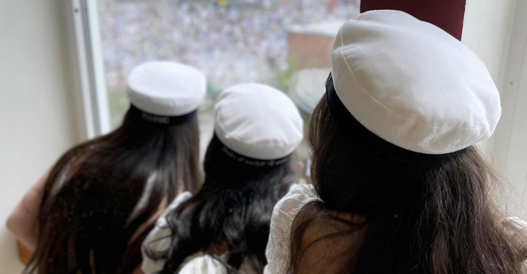 Tre tjejer i studentmössa, bild tagen bakifrån, tittar ut genom fönster.